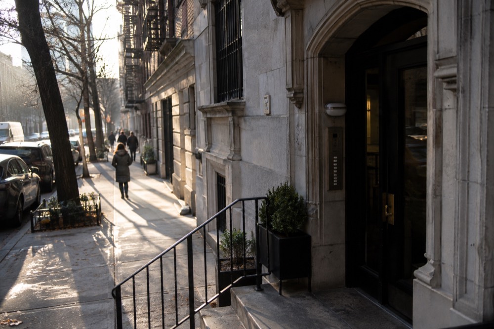A residential apartment building entrance on a New York street with a row of door buzzers beside the door