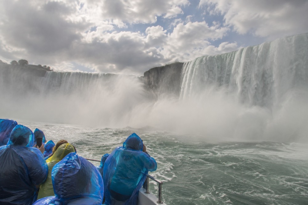 Mist rising over Niagara Falls as visitors in rain ponchos watch from a boat near the base of the falls