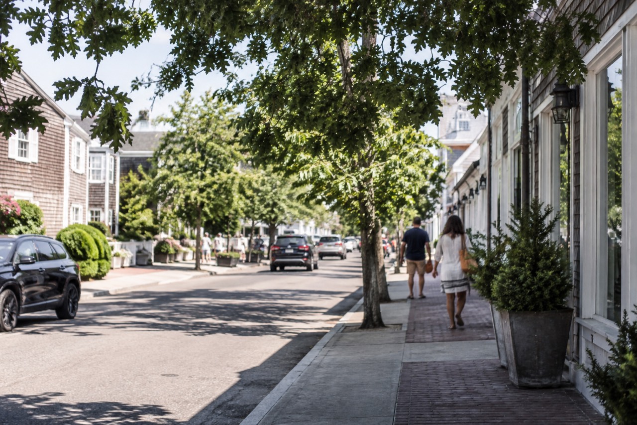 A quiet Hamptons village street with shingled houses and storefronts on a sunny afternoon