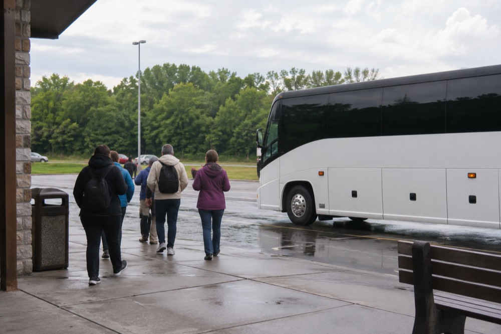 A coach parked at a rest stop beside a highway with trees beyond the lot