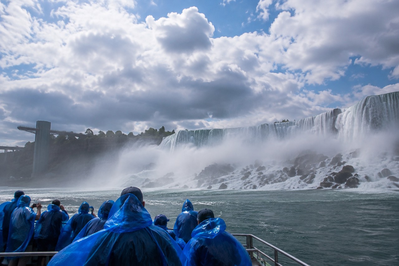A tour boat in blue rain ponchos approaching the base of a wide waterfall with mist rising over the gorge