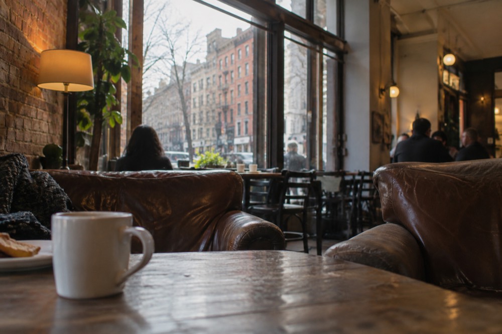 A cozy coffee shop interior in Greenwich Village with a large window, worn couches and warm lighting