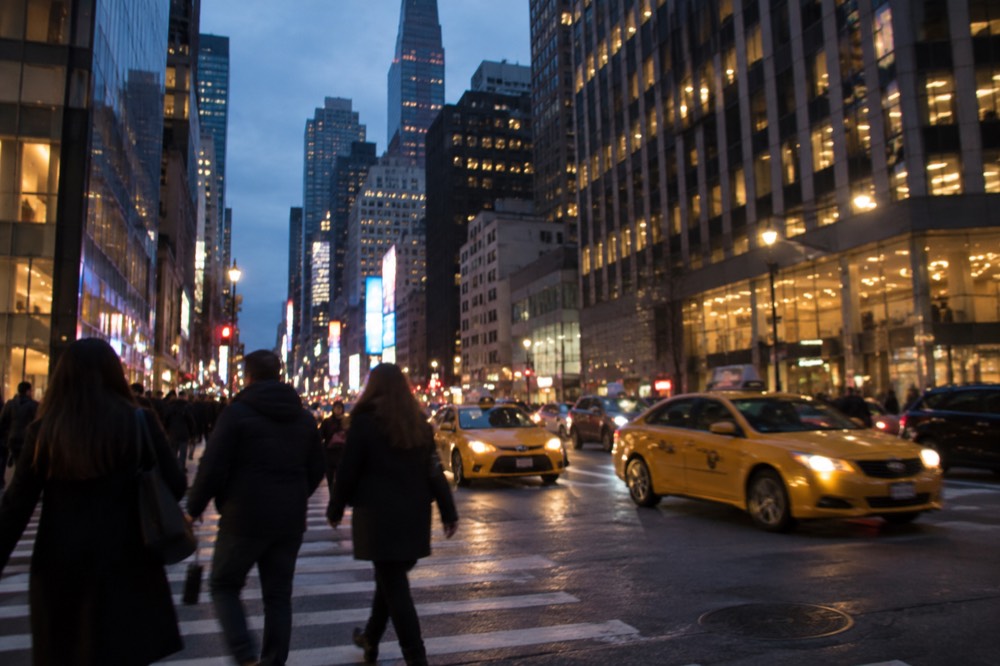 Pedestrians crossing a wide Midtown avenue with yellow taxis in the evening light