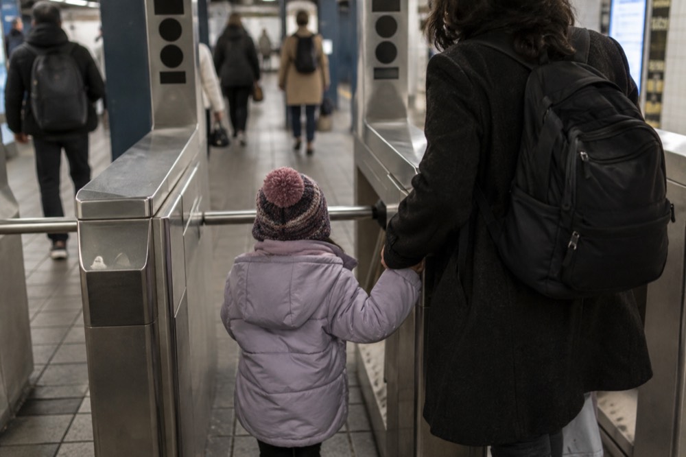 Child holding a parent's hand while stepping through a subway turnstile in a tiled station