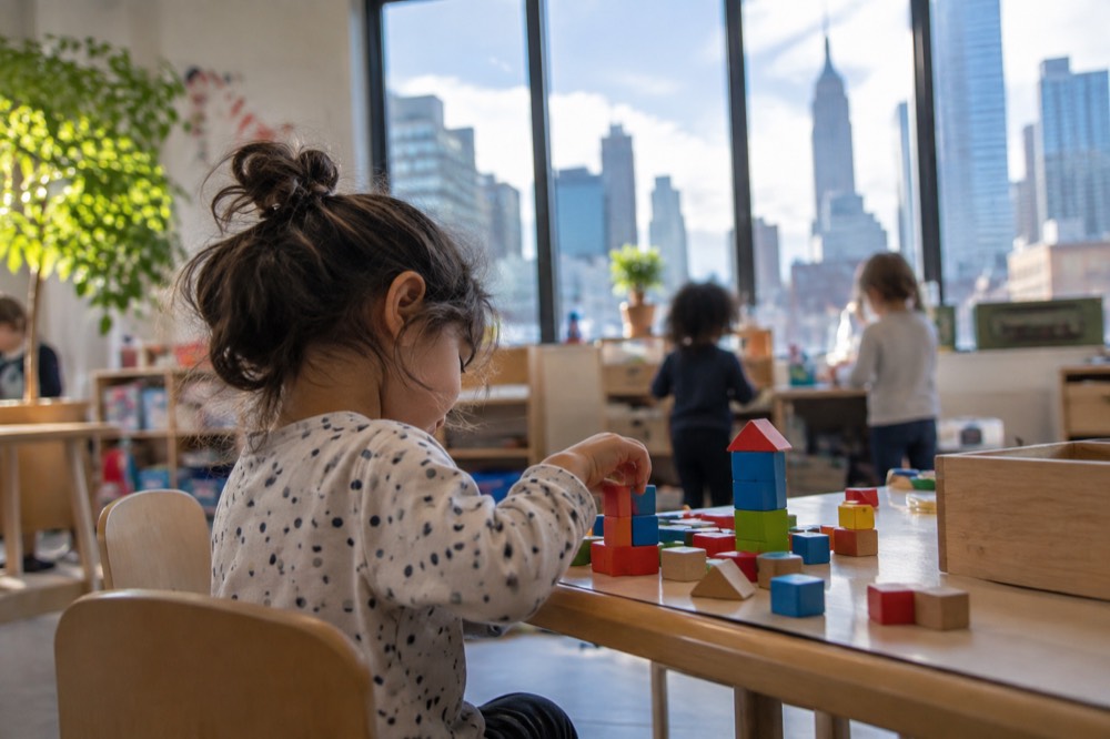 A young child sitting at a small classroom table with building blocks in a bright preschool room