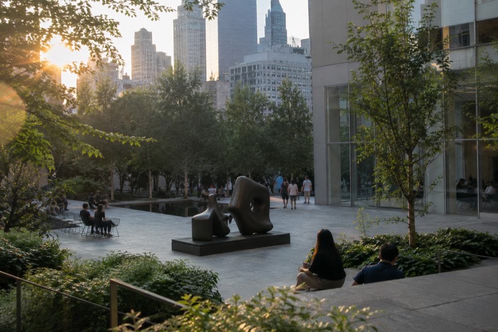 An outdoor sculpture garden with abstract bronze statues, green trees and people sitting on benches beside a modern glass building