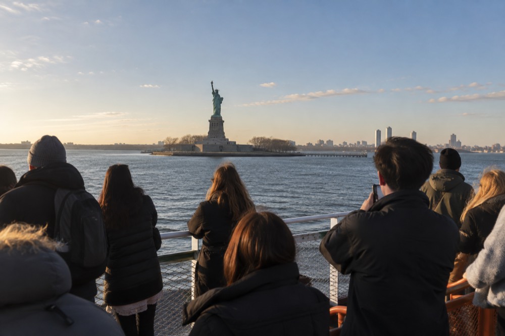 Visitors on a ferry deck looking toward the Statue of Liberty across New York Harbor on a clear day.
