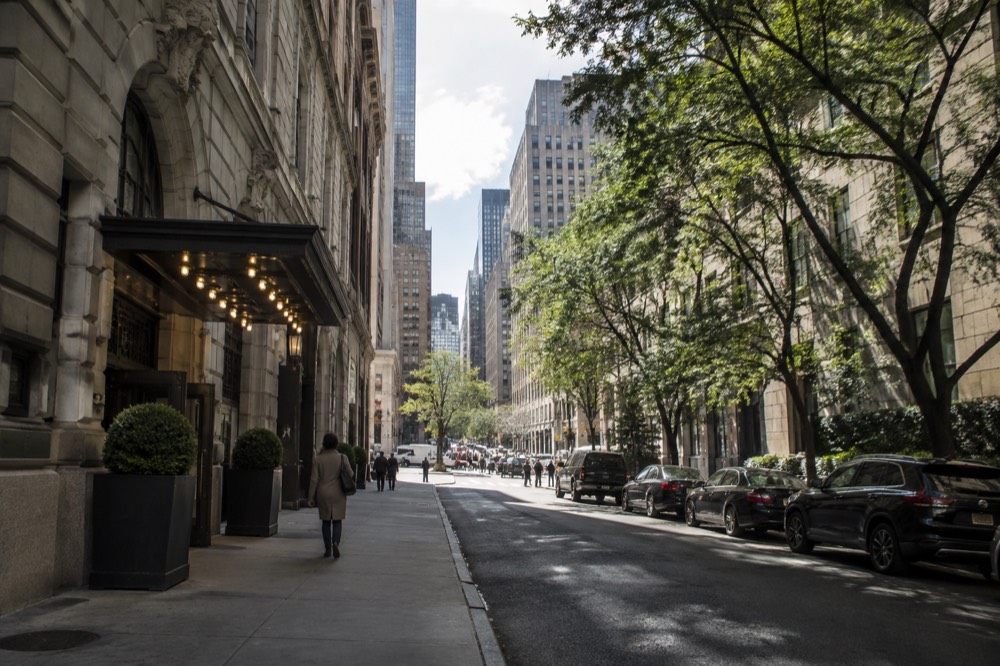 A quiet tree-lined Midtown side street with hotel awnings, contrasting with the bright crowded avenue at the corner