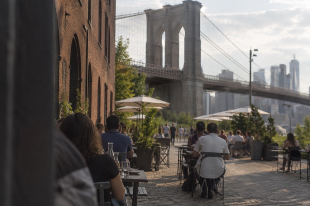 Diners at outdoor tables on a cobblestone street with the Brooklyn Bridge towers overhead