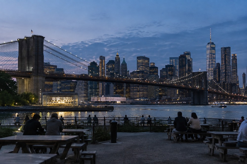 The Brooklyn Bridge towers seen from a waterfront pier with picnic tables and the Manhattan skyline behind