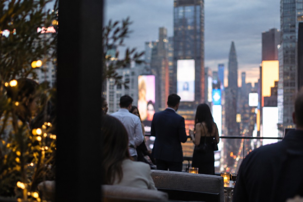 A rooftop bar terrace at dusk with the glowing billboards of Times Square spread out below.