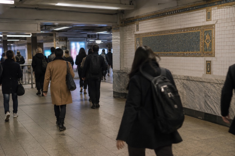 Commuters crossing a tiled New York subway mezzanine with mosaic station name plaques