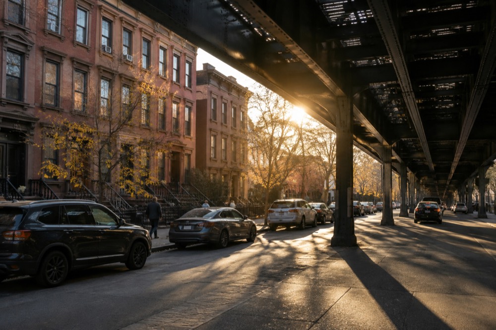 A Brooklyn street corner with row houses and an elevated subway track overhead in late afternoon light