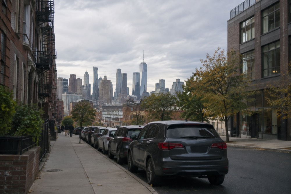 A quiet side street in Brooklyn with parked cars along the curb and the Manhattan skyline in the distance