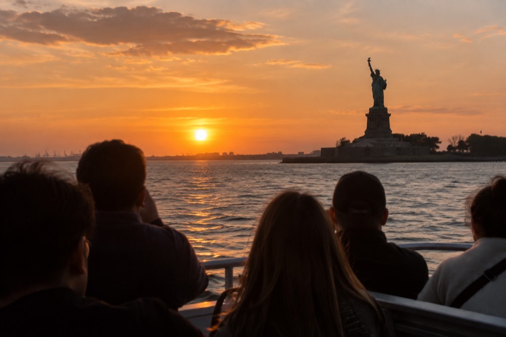 A sightseeing boat on New York Harbor at sunset, with the Statue of Liberty silhouetted against an orange sky