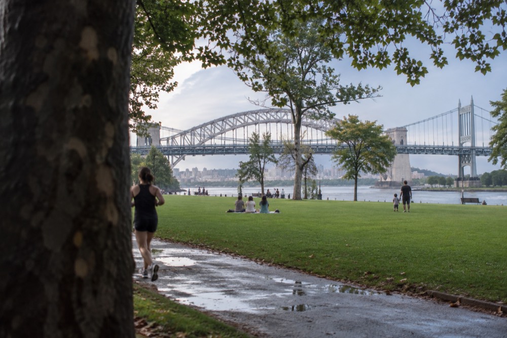 Families and joggers on the lawns of Astoria Park with the East River and bridges in the background