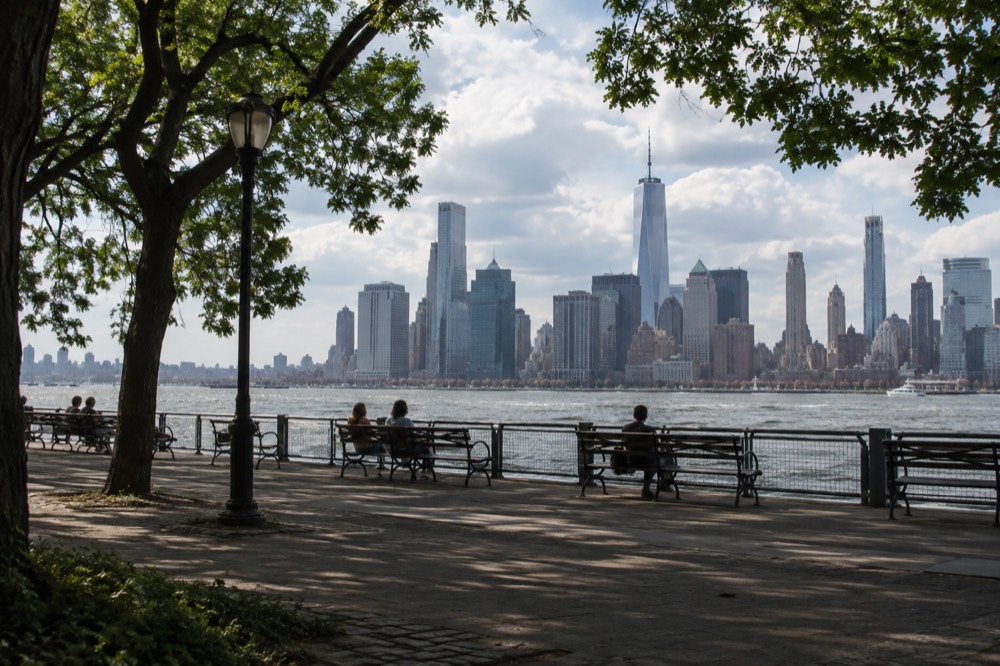 A tree-lined promenade with benches facing the Lower Manhattan skyline across the East River