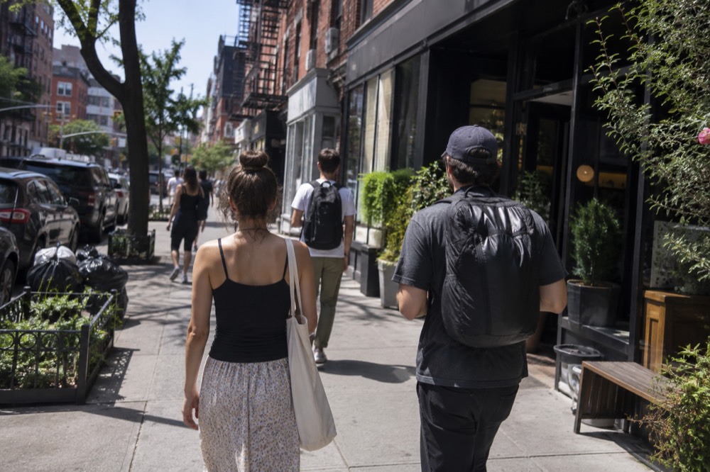 People walking along a narrow East Village sidewalk past storefronts and hanging signage
