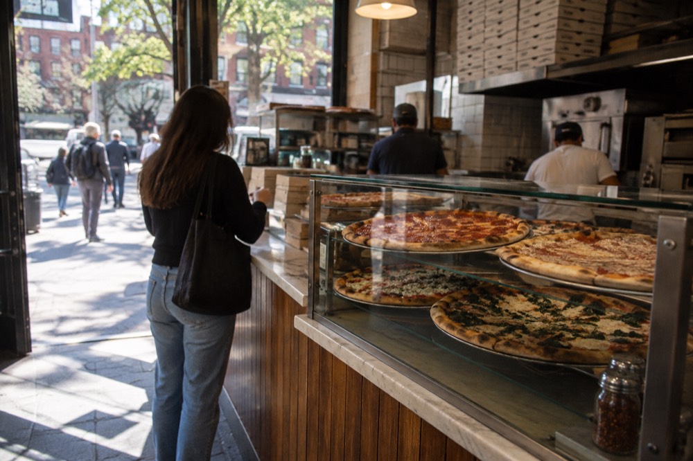 Counter of a Village pizza shop with pies on display and a customer ordering a slice
