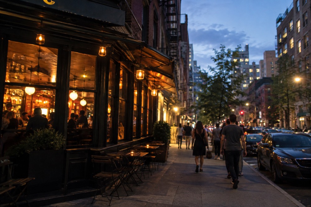 A evening street scene on the Lower East Side with lit bar and restaurant signs and people outside
