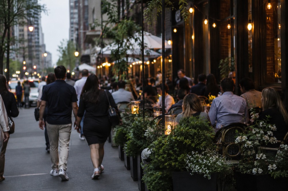 A busy restaurant sidewalk with small tables and string lights in the evening