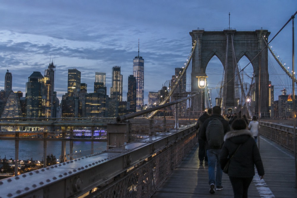 Pedestrians crossing the Brooklyn Bridge wooden promenade with suspension cables rising overhead