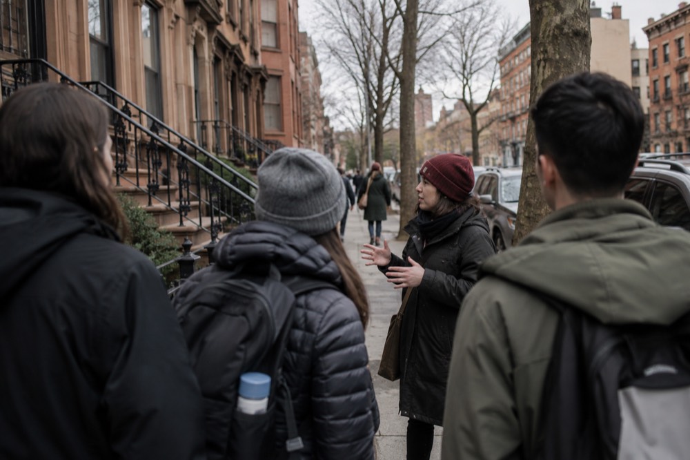 A guide speaking to a small group of visitors on a Brooklyn sidewalk