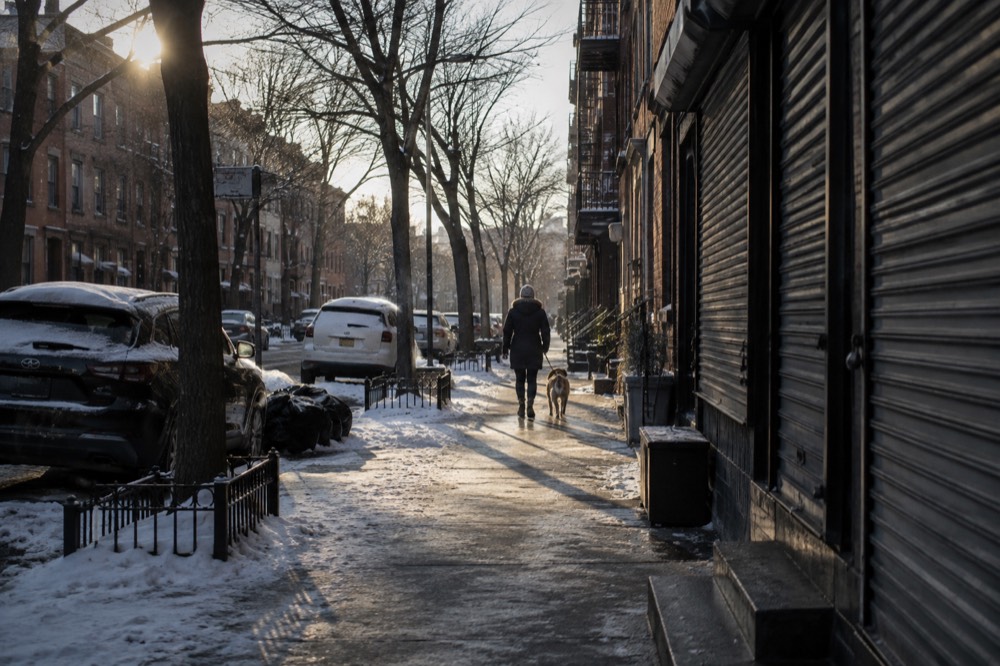 Snow-dusted street with closed storefronts and one person walking a dog