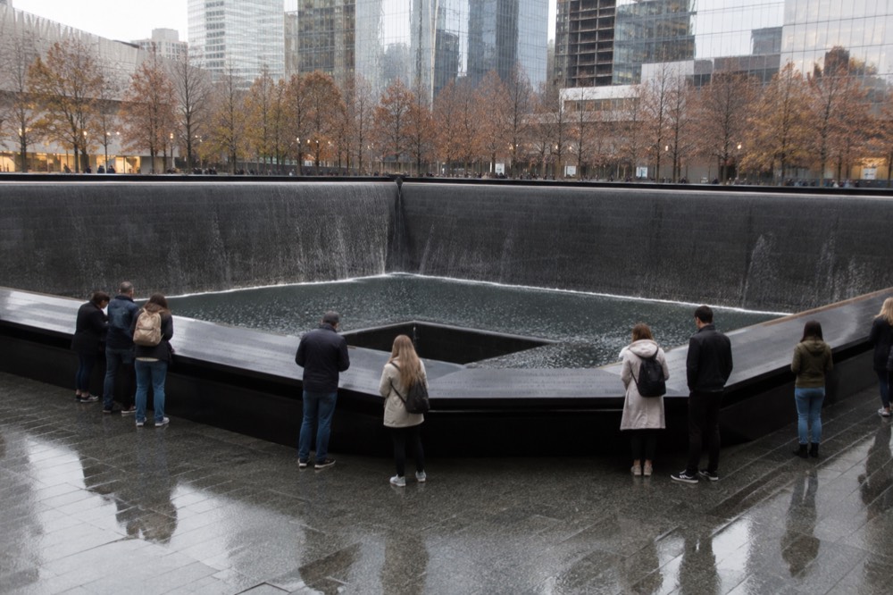 Visitors standing quietly at the edge of a memorial pool, reading names cut into the bronze parapet