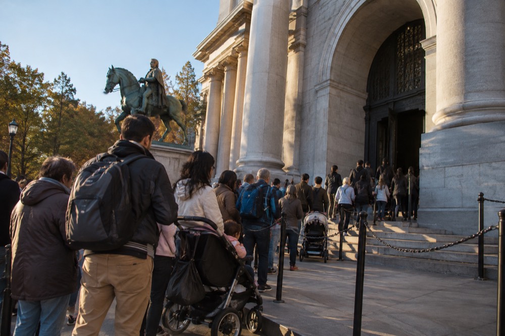 A long queue of visitors with strollers and backpacks waiting outside a museum entrance on a sunny morning