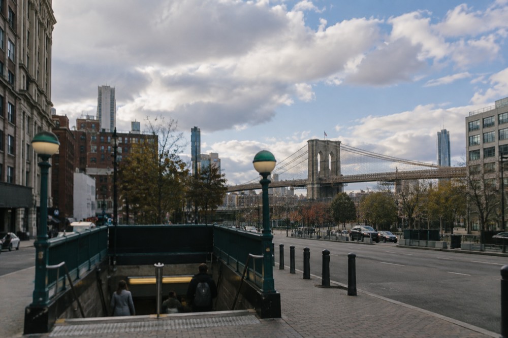 A subway entrance in lower Manhattan with the Brooklyn Bridge visible behind the rooftops
