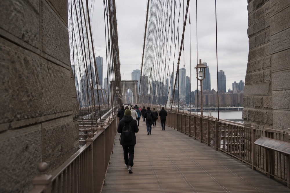 The wooden planks of the Brooklyn Bridge promenade narrowing between the stone tower and steel cables