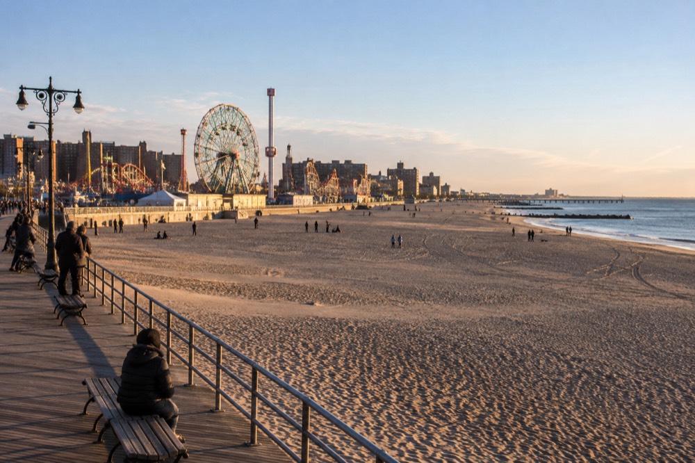 A wide sandy beach with a wooden boardwalk and amusement park rides rising against the sky