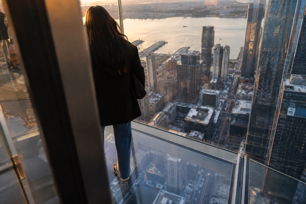 A person standing on a transparent glass floor panel looking down at streets far below