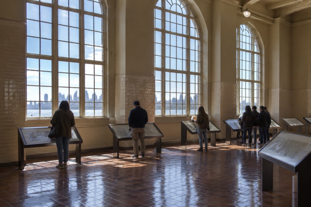 Visitors reading information panels inside the Ellis Island museum registry room under tall windows