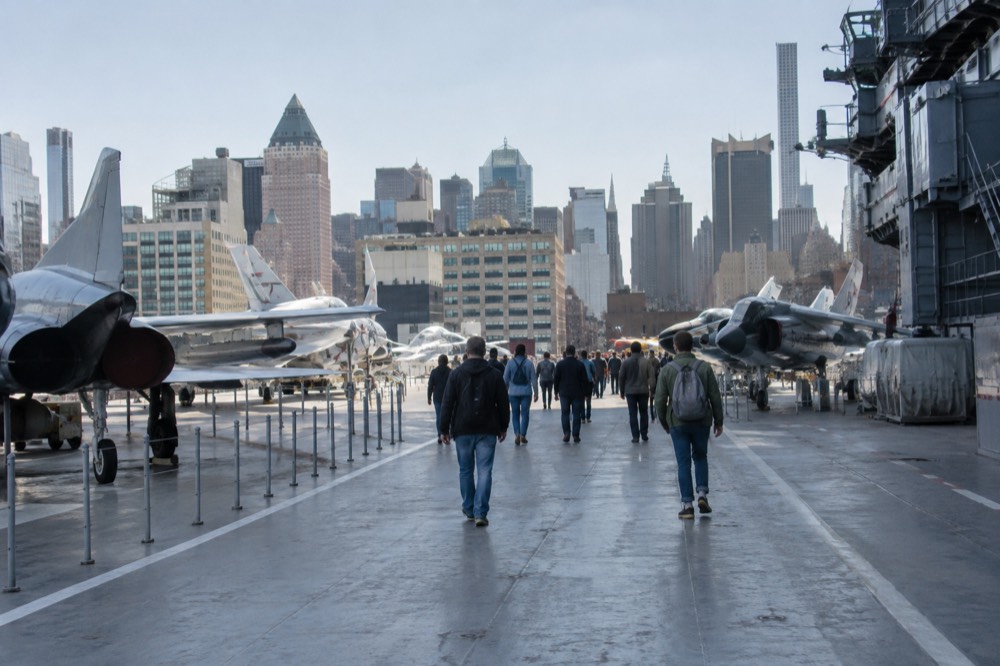 Visitors walking among fighter jets parked on the carrier's flight deck under a clear sky