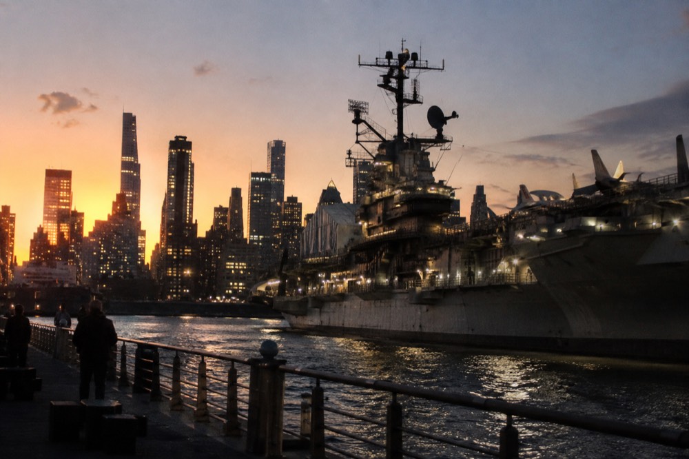 The Intrepid moored at dusk, its flight deck lights on and the Manhattan skyline glowing behind the ship