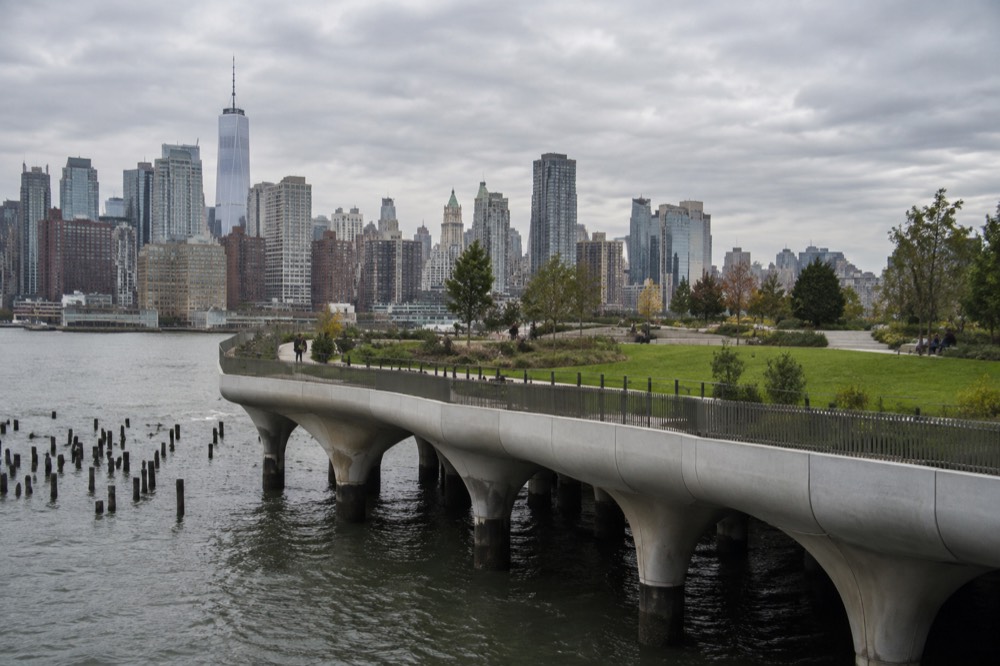 A curved elevated park on stilts above the Hudson River with green lawns and the Manhattan skyline behind it