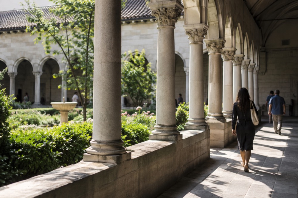 A stone cloister walkway with arched columns opening onto a planted garden