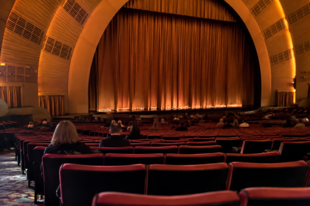Rows of red seats in a large theater auditorium curving toward a wide stage with the curtain down