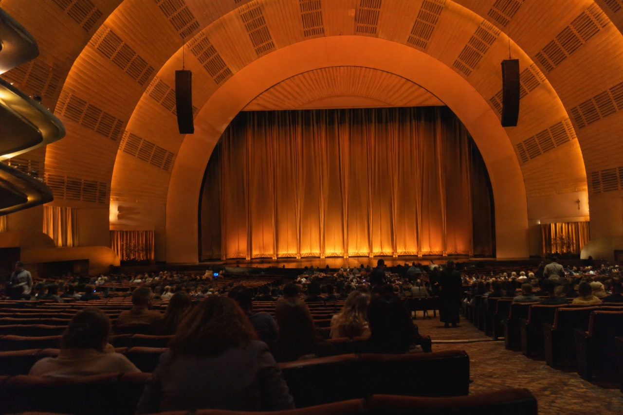 The auditorium of Radio City Music Hall seen from the rear of the orchestra seating, with the stage curtain closed and rows of seats curving toward it