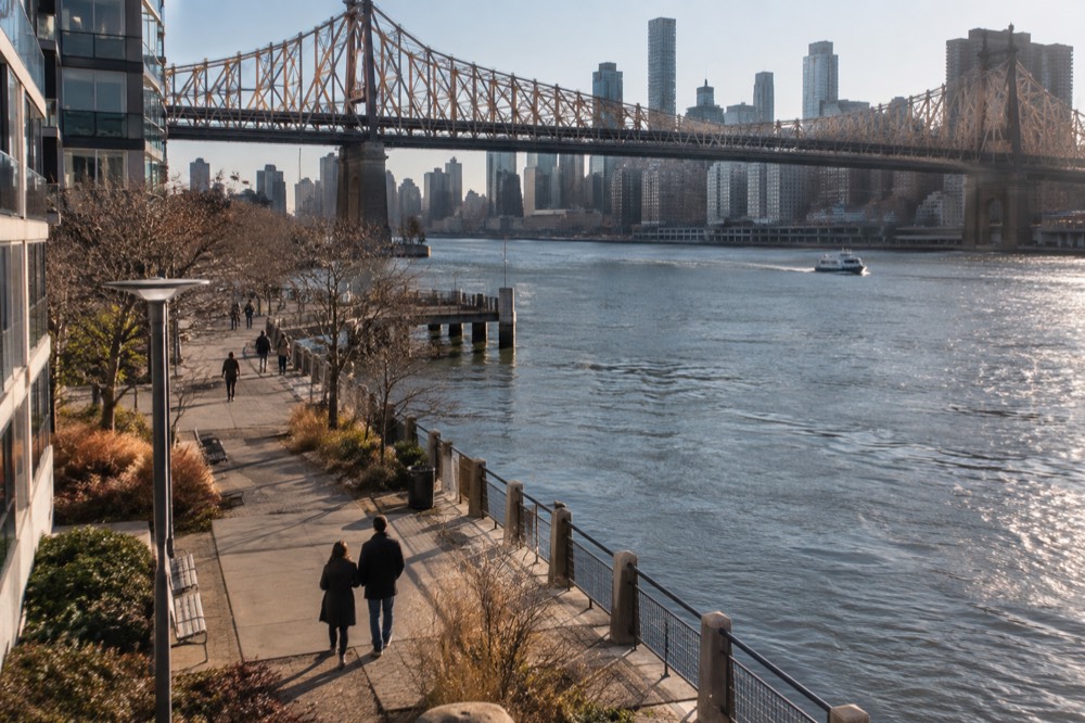 A quiet stretch of the Roosevelt Island promenade in late afternoon light with the Queensboro Bridge above