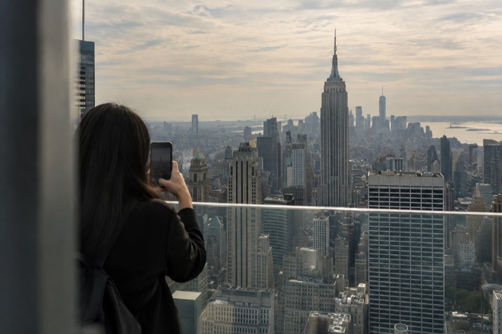 A visitor photographing the skyline with a phone from behind a glass railing high above Midtown office towers