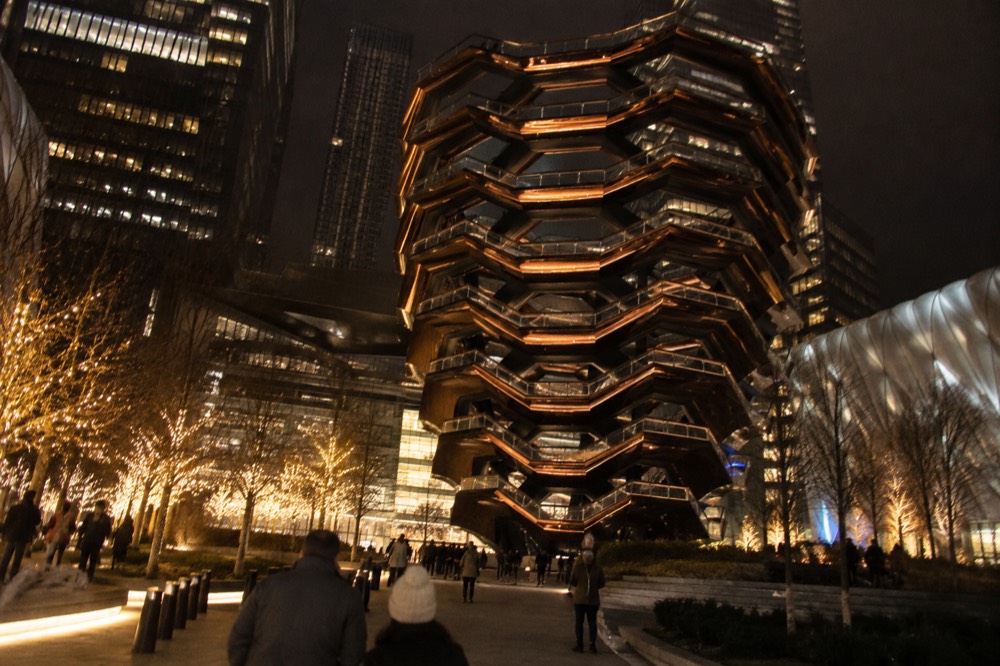 Vessel lit up at night with its staircases glowing against a dark sky above the Hudson Yards plaza