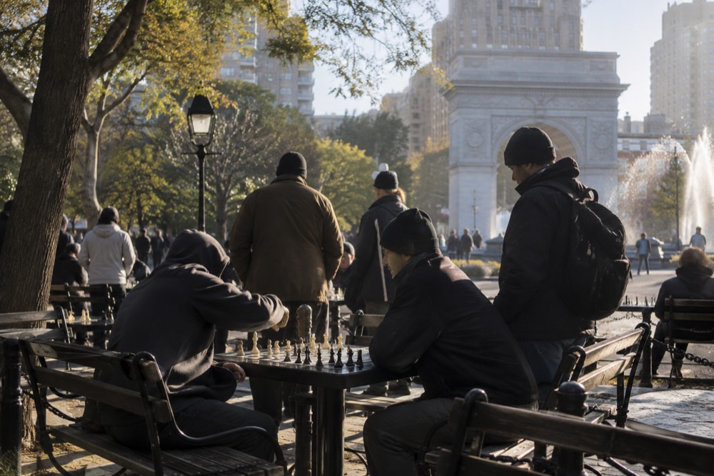 Chess players mid-game at the outdoor tables in Washington Square Park with onlookers standing behind