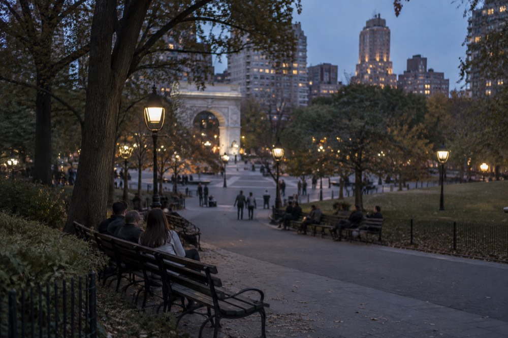 Visitors sitting on benches under trees beside a paved path in Washington Square Park