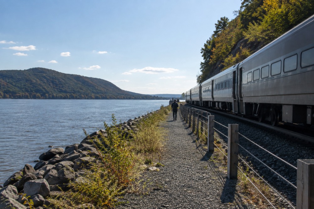 A train travelling along tracks beside a wide river with wooded hills on the far bank
