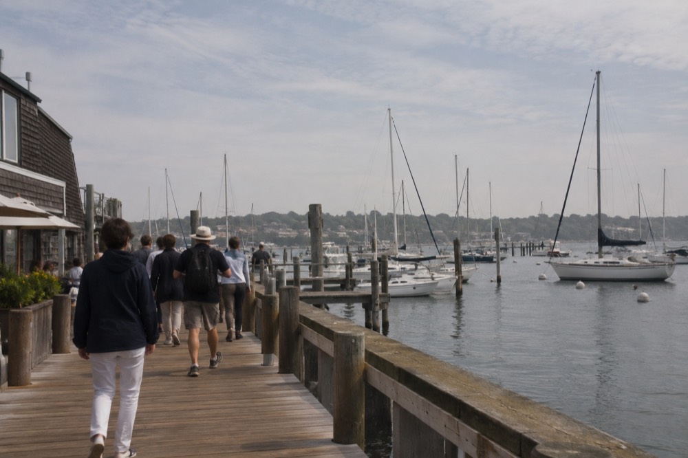 A guided tour group walking along a wooden boardwalk beside a harbor full of sailing boats