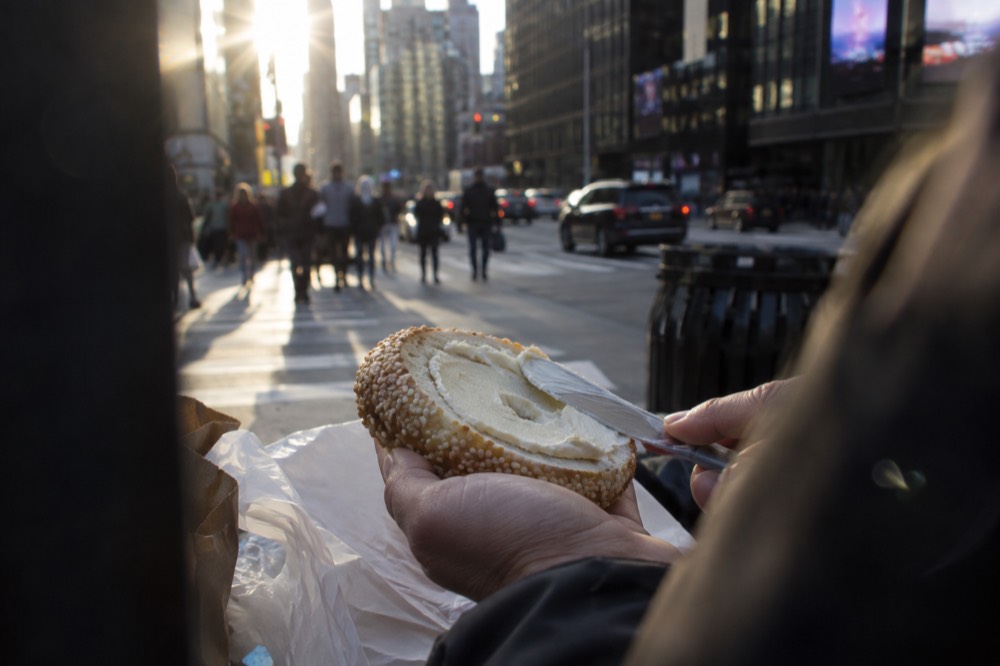 A close view of a sesame bagel with cream cheese being spread with a knife