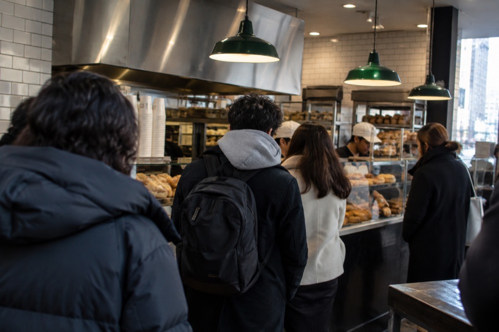 A busy bagel shop counter with customers waiting in a line inside a small shop